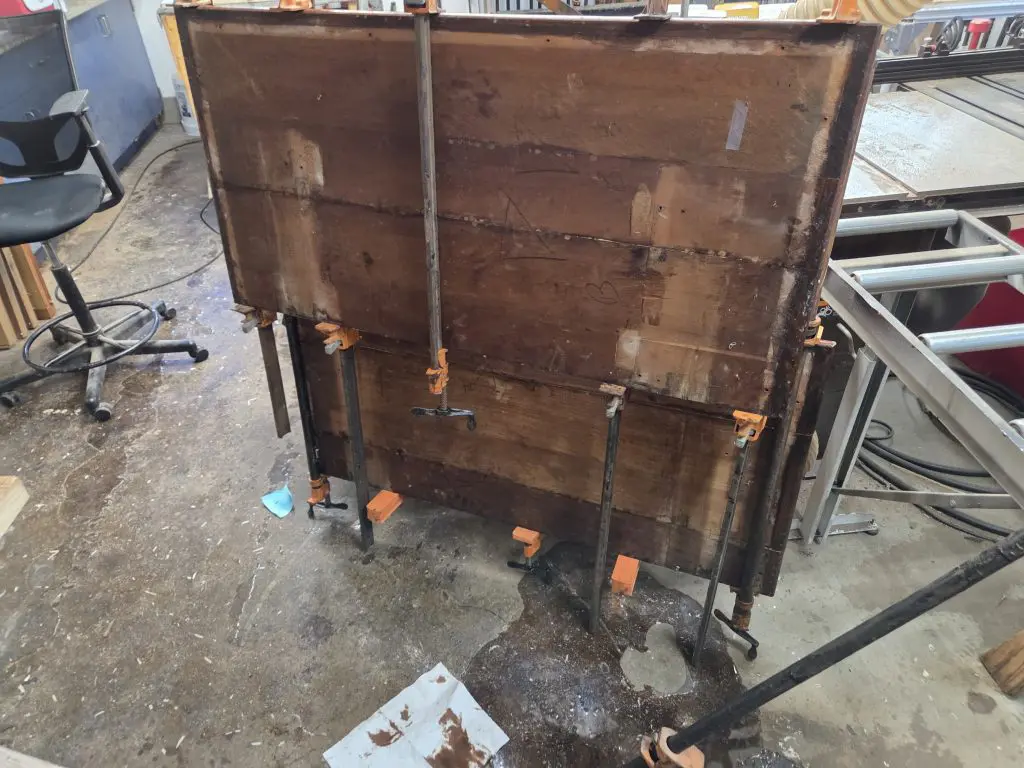 20250911_135322 - Four Fields Furniture A large, weathered wooden crate mounted on adjustable metal stands with clamps in a cluttered workshop. Nearby chair, cords, and tools are visible on the concrete floor.