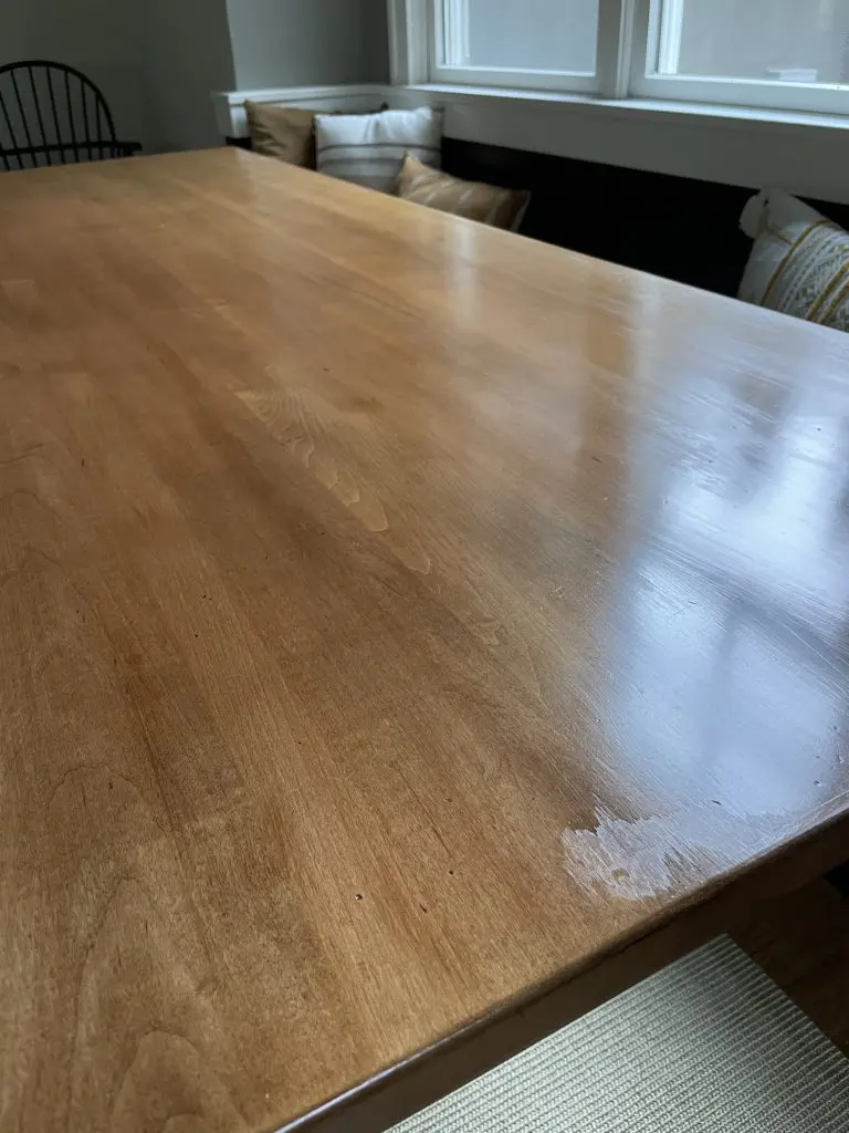IMG_2735 - Four Fields Furniture Wooden dining table with visible scratches and grain; cushions on a bench and a chair in the background by a window.
