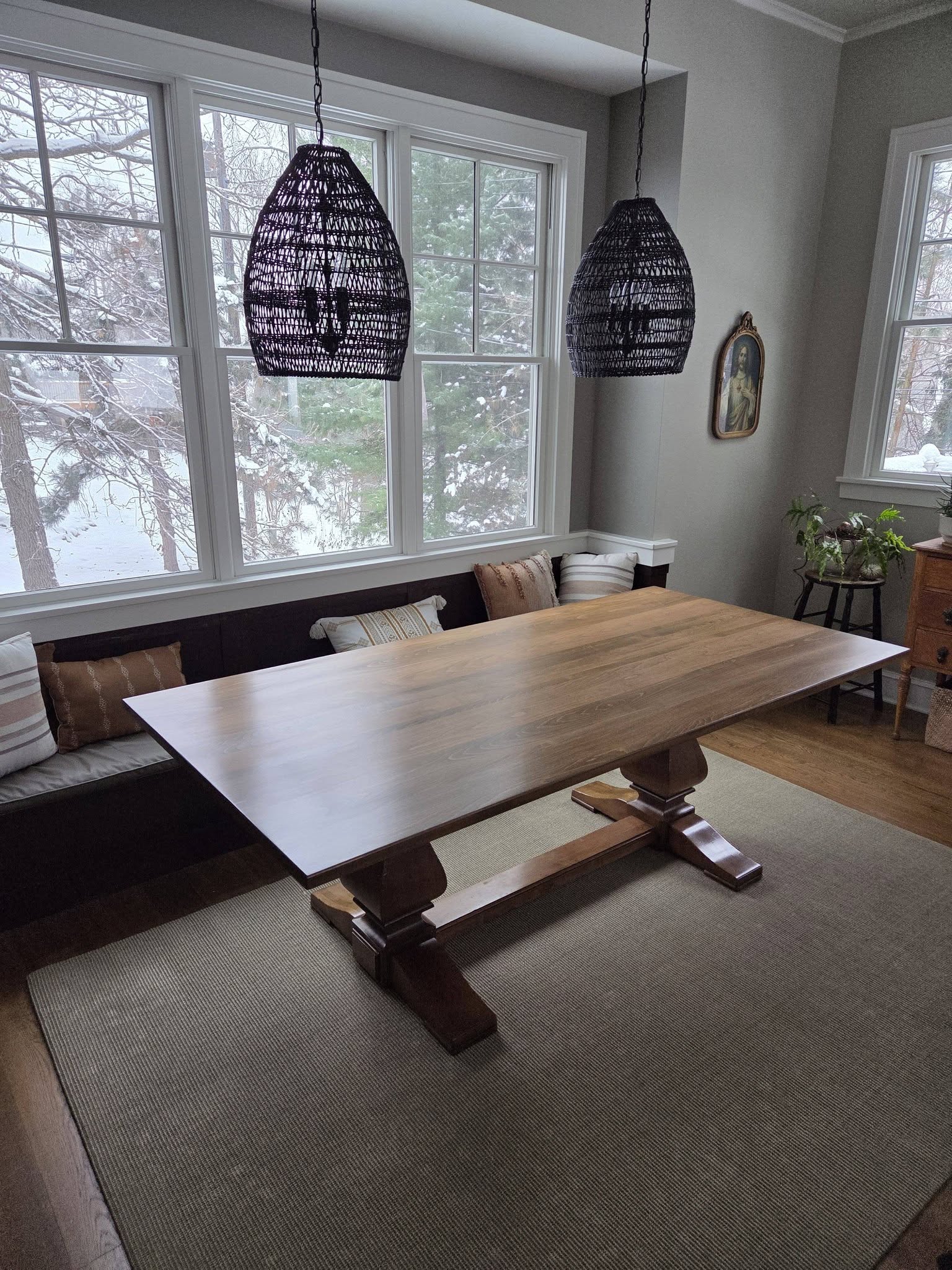 598258040_1407985934654053_5157564628800985719_n - Four Fields Furniture A dining area with a large wooden table, dark bench seating, and two woven pendant lights hanging from the ceiling; snowy windows outside and a small plant nook nearby.